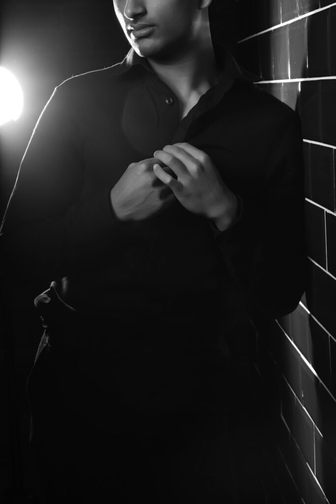 Man in black shirt adjusting collar against tiled wall with spotlight.
