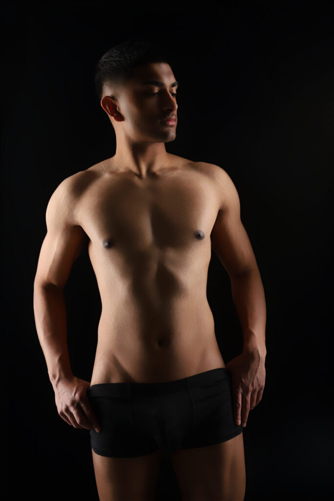Man posing in black underwear against dark background.